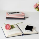 A stylish home office setup with open book, smartphone, and flowers on a white desk.